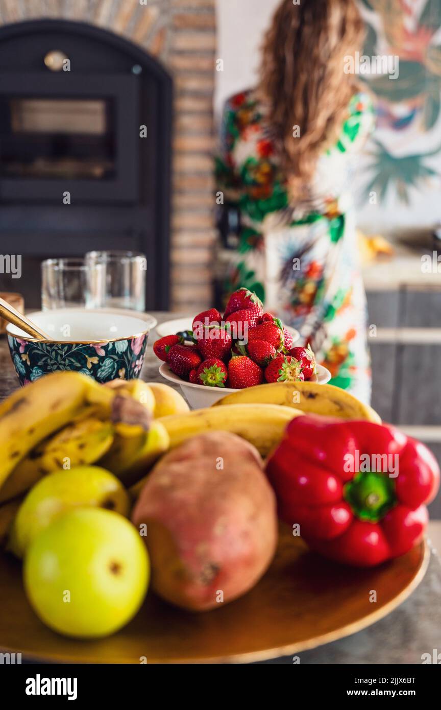 Set von bunten reifen Früchten und Gemüse auf dem Tisch neben Obst und Geschirr in der hellen Küche mit gesichtslosen Frau platziert Stockfoto