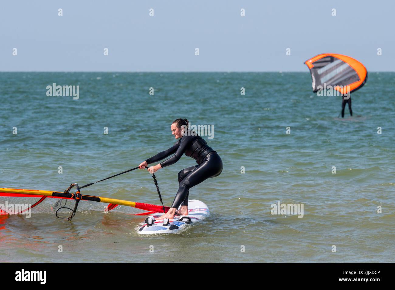 Mädchen / weibliche Freizeit Windsurfer in schwarzem Neoprenanzug üben klassischen Windsurfen und heben Segel auf See Stockfoto