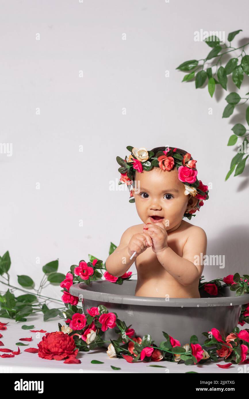 Schöne Latina Baby Mädchen mit brauner Haut, in einem grauen Eimer, umgeben von Blumen und Rosen und rot, mit einem weißen Hintergrund. Kleines Mädchen suckin Stockfoto
