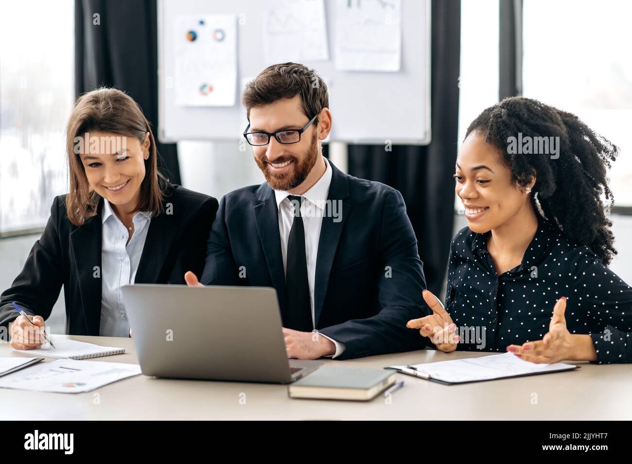 Multirassische Kollegen denken gemeinsam an Strategien, sitzen im modernen Büro, studieren Dokumente, benutzen Laptops, diskutieren, lächeln. Erfolgreiche kreative Geschäftspartner, die an einem Projekt arbeiten Stockfoto