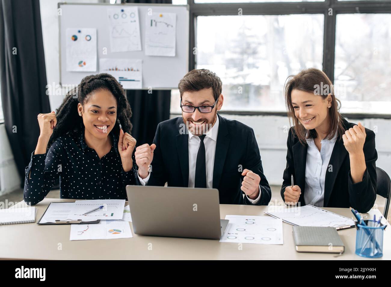 Fröhliche kreative Geschäftspartner arbeiten im Büro zusammen. Multirassische Kollegen, die im modernen Büro sitzen und einen Laptop benutzen, sind glücklich, gute Sache zu bekommen, großen Gewinn. Das Konzept von Sieg, Triumph Stockfoto