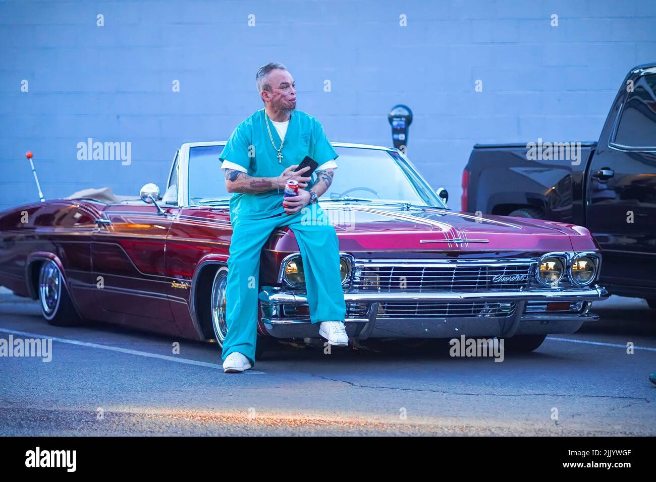 Ein Mann im Hip-Hop-Kleidungsstil lehnt sich an ein klassisches Chevrolet Impala-rotes Auto an Stockfoto