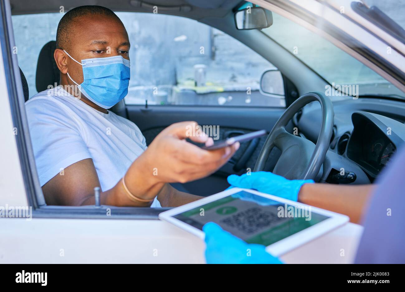 Covid fährt durch einen Checkpoint-Mann, der QR-Code auf einem Tablet scannt, während sein Telefon im Auto unterwegs ist. Mann, der mit einer Gesichtsmaske auf ein Coronavirus fährt Stockfoto