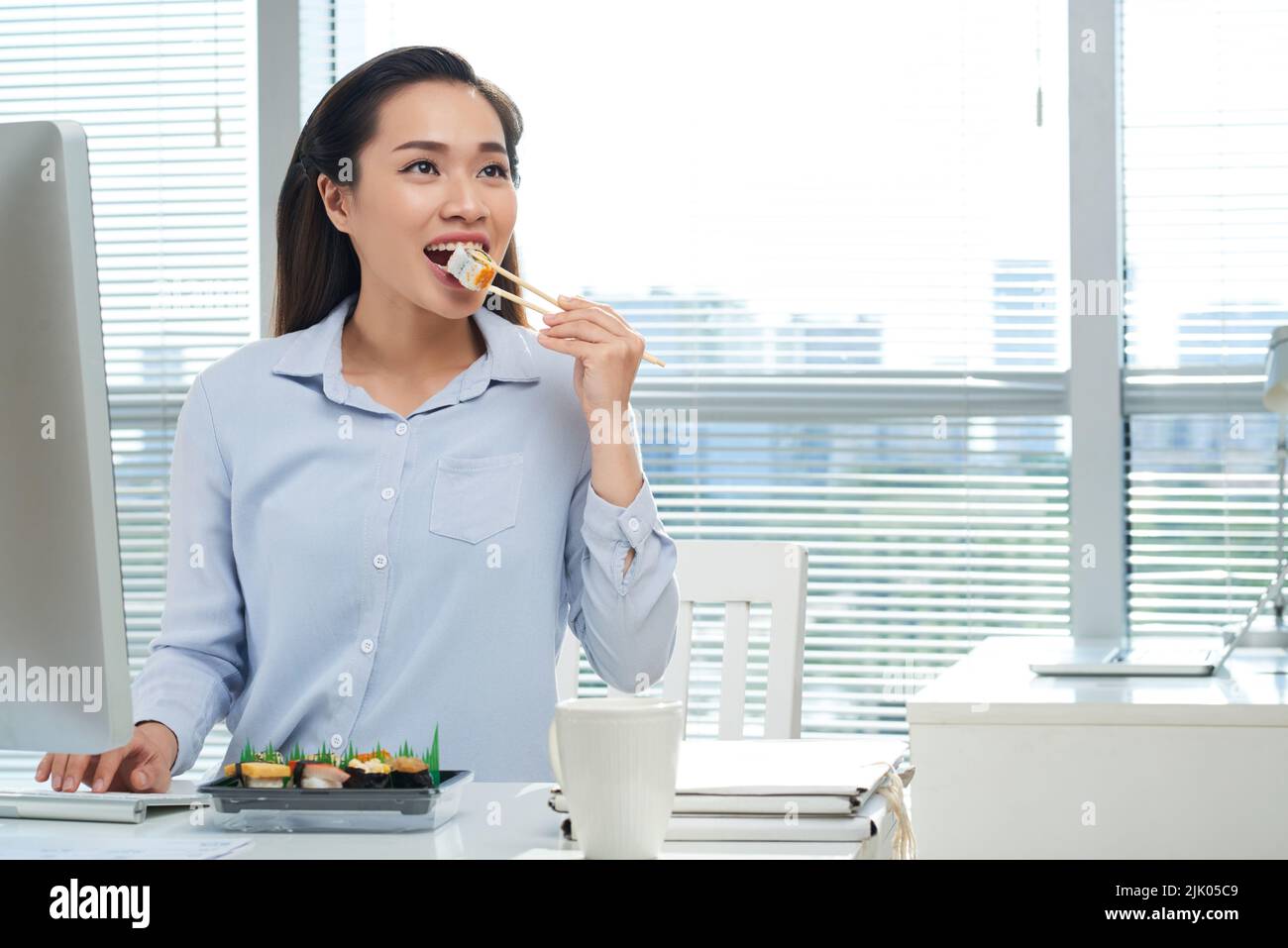 Porträt eines attraktiven asiatischen Managers, der am Schreibtisch sitzt und appetitliches Sushi isst, während er eine kurze Pause von der Arbeit macht, Panoramafenster im Hintergrund Stockfoto