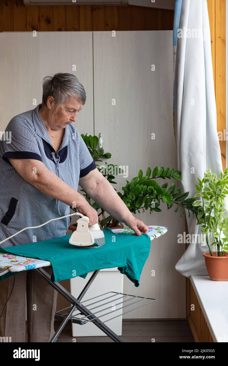 Ältere Dame beim Bügeln zu Hause. Ältere Frau zu Hause kümmert sich um Kleidung. Häusliche Arbeit mit Glück, selektiver Fokus gemacht. Stockfoto