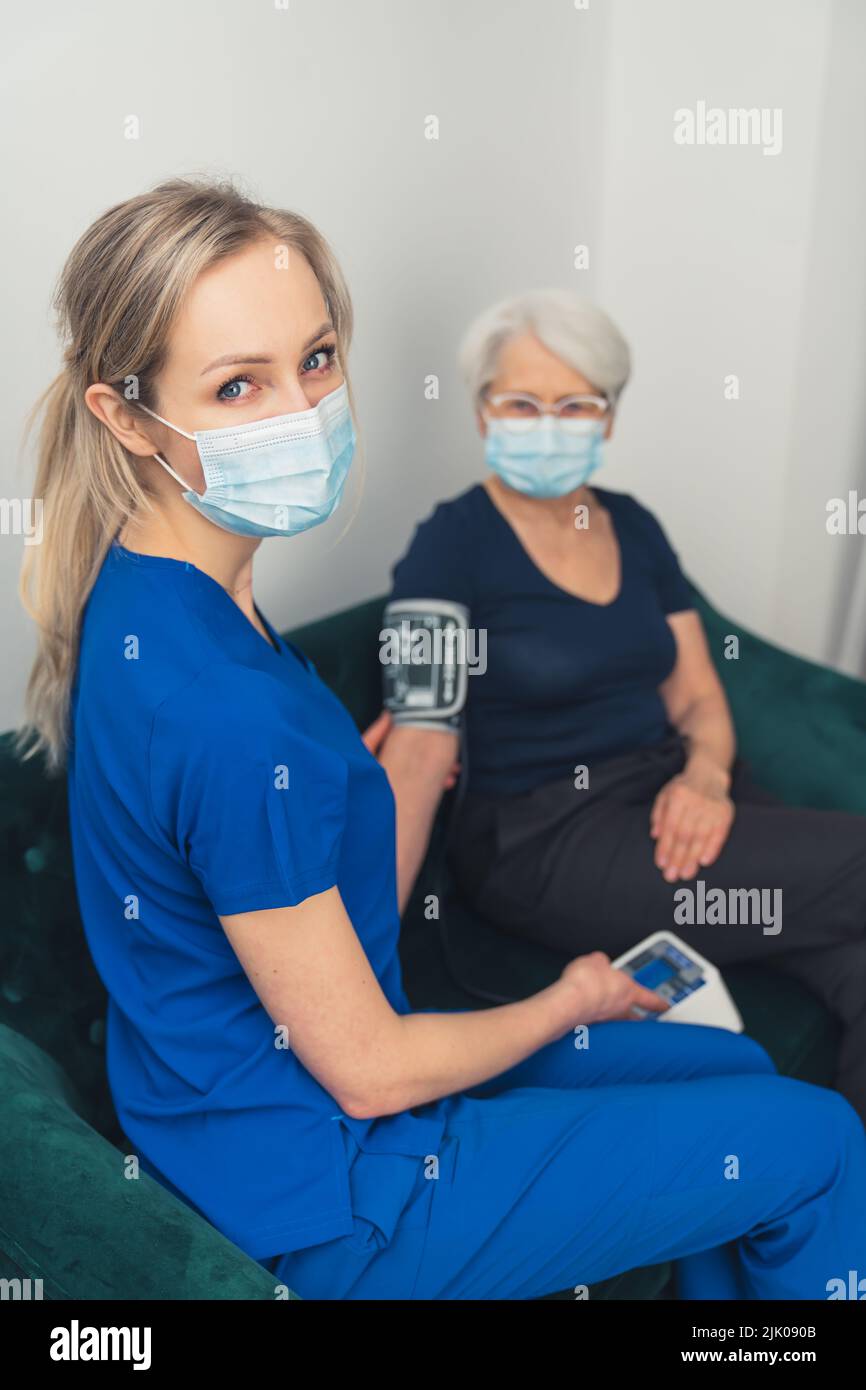 Eine junge, attraktive Krankenschwester mit blauen Augen und langen blonden Haaren in dunkelblauer Uniform, die den arteriellen Blutdruck einer älteren Frau mit einer Gesichtsmaske während einer Pandemie misst. Hochwertige Fotos Stockfoto