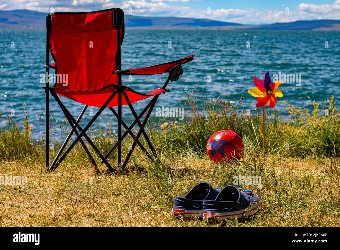 Roter Campingstuhl am Strand an einem Sommertag auf dem Hintergrund des Berges, das Meer . Hausschuhe, Ball und Wetterfahne. Stockfoto
