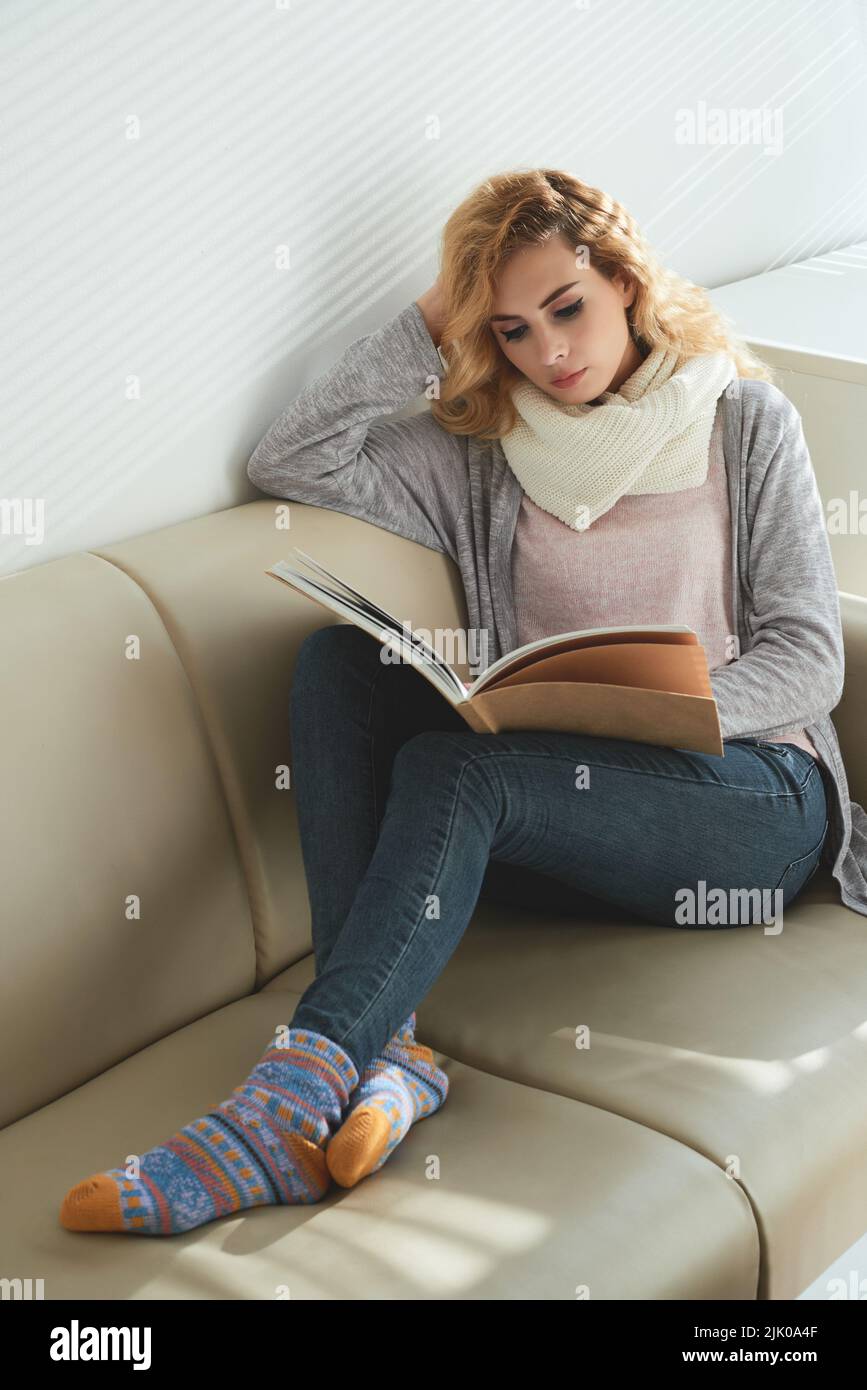 Schöne junge Frau in warmen Kleidern, die sich mit einem Buch auf dem Sofa ausruht Stockfoto