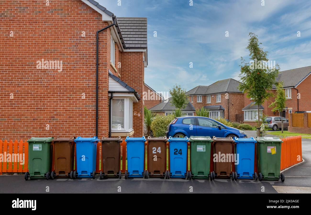 Eine Reihe farbiger Mülltonnen zum Recycling von Kunststoff-, Papier- und Gartenabfällen, neben einem orangefarbenen Zaun und vor einem modernen Haus. Stockfoto