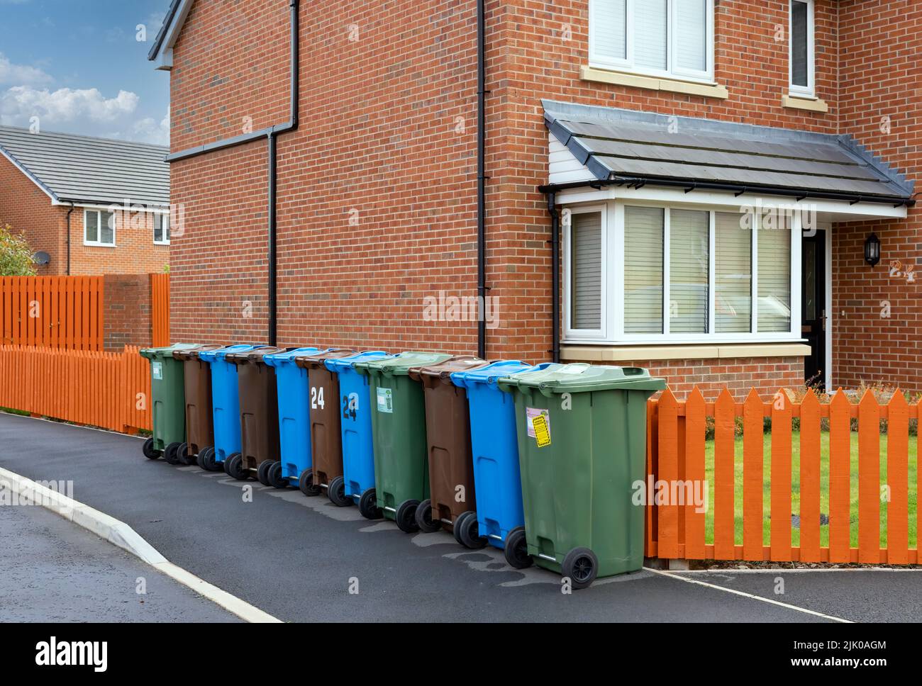 Eine Reihe farbiger Mülltonnen zum Recycling von Kunststoff-, Papier- und Gartenabfällen, neben einem orangefarbenen Zaun und vor einem modernen Haus. Stockfoto