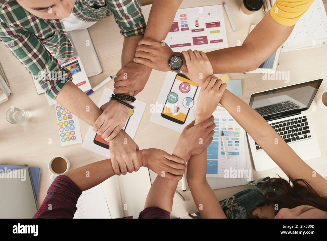 Junge Menschen halten sich gegenseitig die Hände in einer Kette über dem Tisch mit Dokumenten für mobile Apps Stockfoto