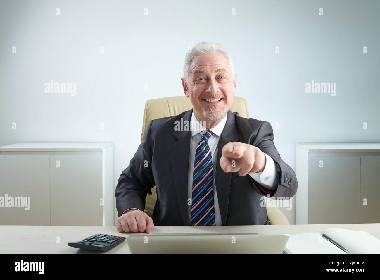 Ein fröhlicher Geschäftsmann im Alter, der in einem klassischen Anzug auf die Kamera zeigt, während er in einem geräumigen Büro von der Arbeit abgelenkt ist, in einem Porträt mit Hüfthöhe Stockfoto