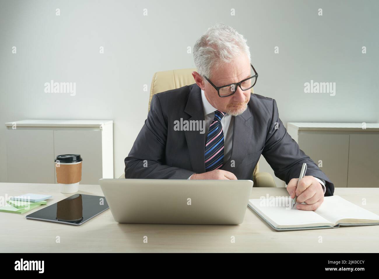 Geballter älterer Geschäftsmann, der in klassischem Anzug und Brillen die notwendigen Notizen macht, während er am Schreibtisch sitzt und sich auf wichtige Verhandlungen vorbereitet, Porträtaufnahme Stockfoto