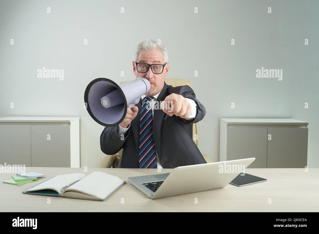 Bärtiger Unternehmer, der einen Anzug mit Megaphon trägt und auf die Kamera zeigt, während er am Schreibtisch eines modernen Büros sitzt und ein Porträt mit Hüfthöhe zeigt Stockfoto