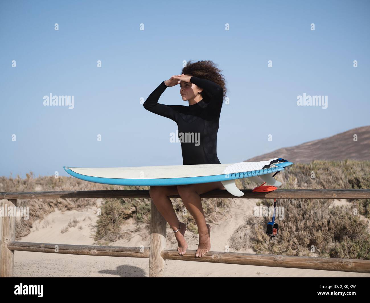 Junge Surferin mit afro Haaren, die die Wellen auf einem Zaun sitzend beobachten Stockfoto