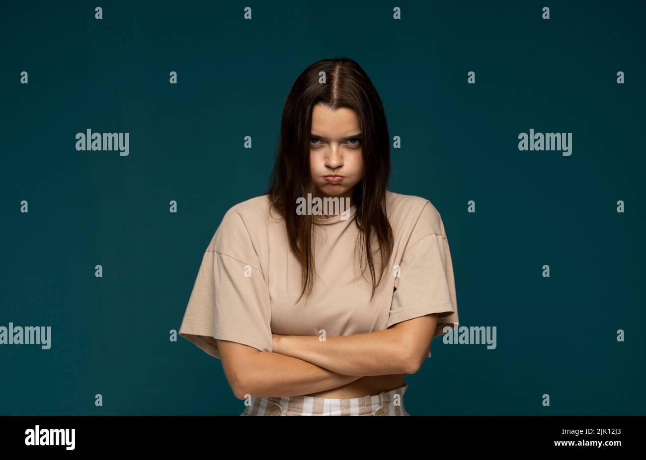 Porträt eines verärgert jungen schönen Mädchen in einem beigen T-Shirt stehen mit geschwollenen Wangen und Arme isoliert auf blauem Hintergrund gefaltet. Emotionen der Wut Stockfoto