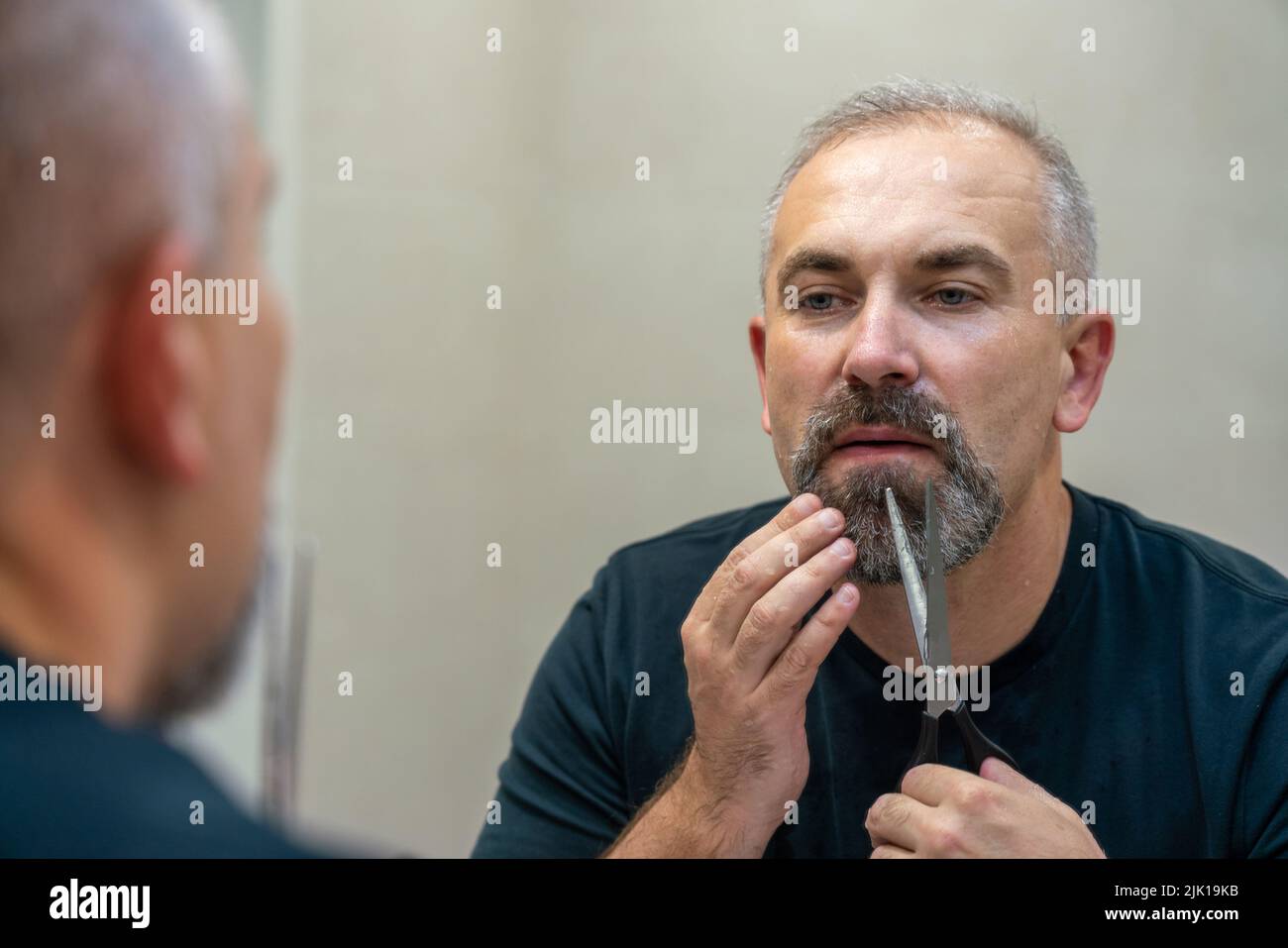 Ein gutaussehender Mann mittleren Alters, der mit einer Schere seinen Bart ein bisschen abschneidet und die Form fixiert Stockfoto