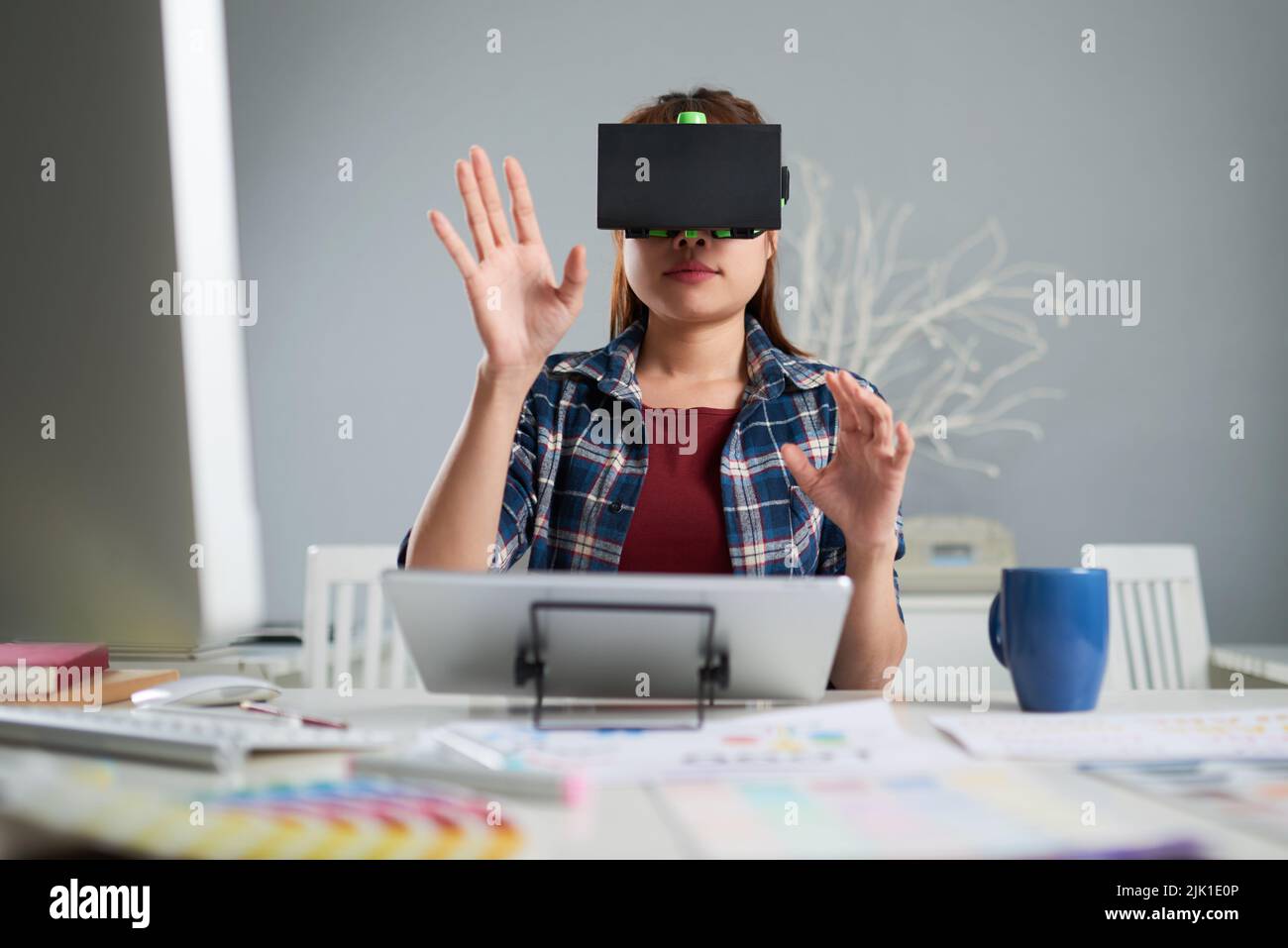 Junge Innenarchitektin in Casualwear, die ein VR-Headset für die Arbeit mit 3D-Visualisierung verwendet, während sie am Schreibtisch eines geräumigen Büros sitzt Stockfoto