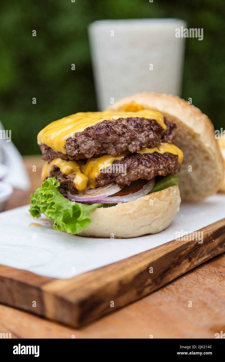 Doppelter Cheeseburger mit Zwiebeln, Tomaten, Salat und Cheddar-Käse auf einem Holzbrett Stockfoto