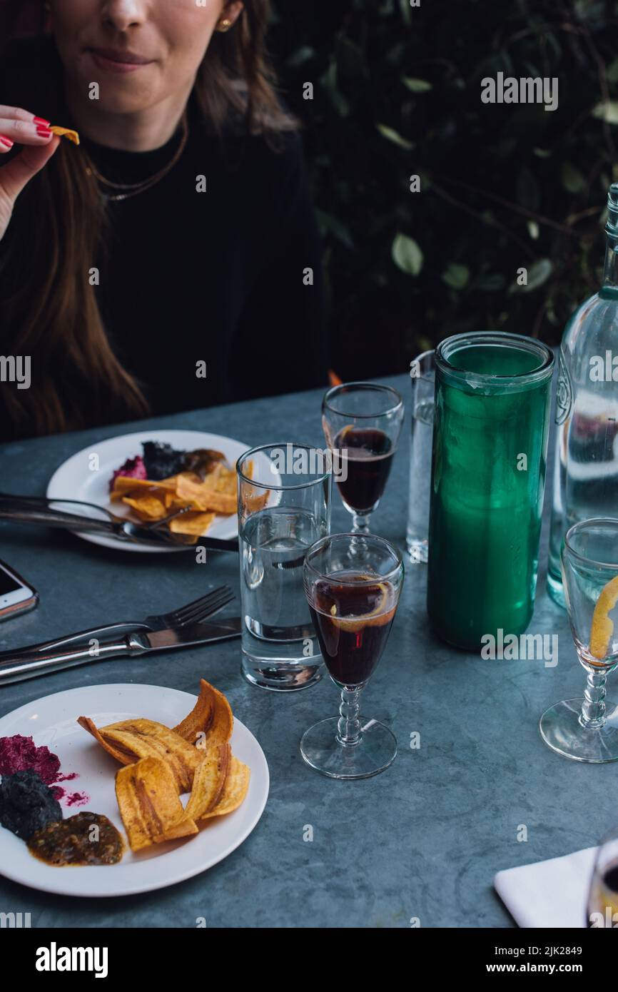 Eine Frau, die im Restaurant im Freien auf einer Metallterrasse mit Weingläsern Plantain Chips und Dips isst Stockfoto