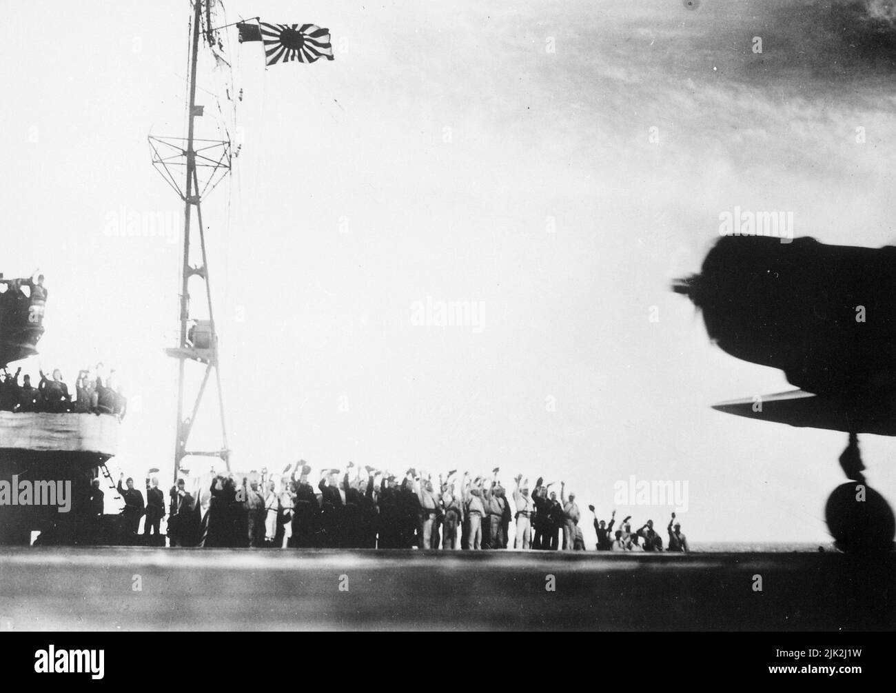 Ein erfasstes japanisches Foto, das vor dem Angriff auf Pearl Harbor an Bord eines japanischen Transporters aufgenommen wurde, 7. Dezember 1941. Es zeigt die Männer, die ihre Arme zum Gruß erheben, mit der Silhouette eines Flugzeugs und der Flagge der aufgehenden Sonne. Der Propeller im Flugzeug dreht sich, so dass dieses Bild möglicherweise aufgenommen wurde, als die Flugzeuge in ihrer berühmten und berüchtigten Mission abreisten. Stockfoto
