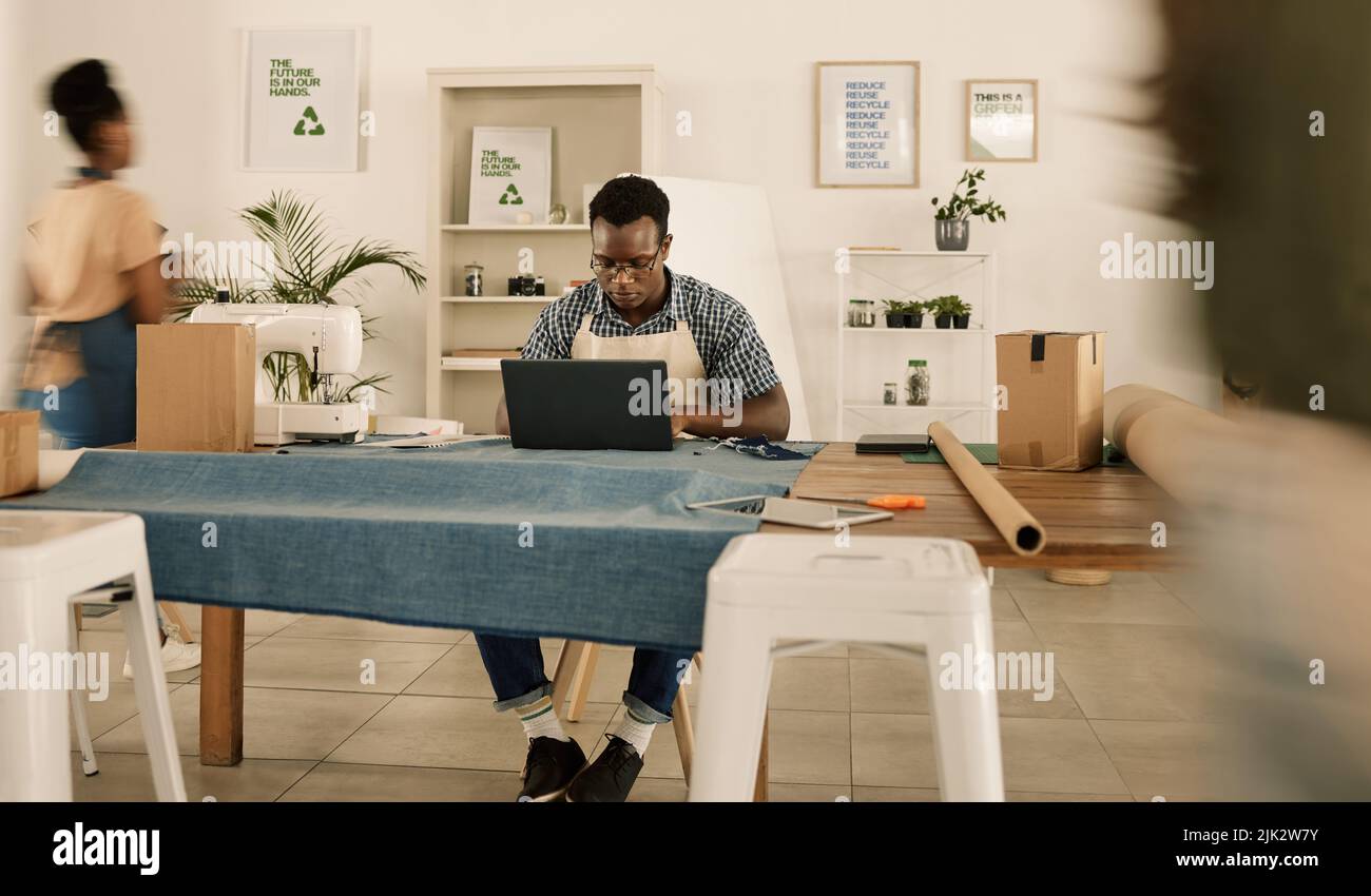 Nachhaltiger Modedesigner, junger Mann und Kreative arbeiten an einem Laptop in einem geschäftigen Bekleidungs-Startup, einer Textilwerkstatt und einem Style-Studio. Fabrikarbeiter Stockfoto