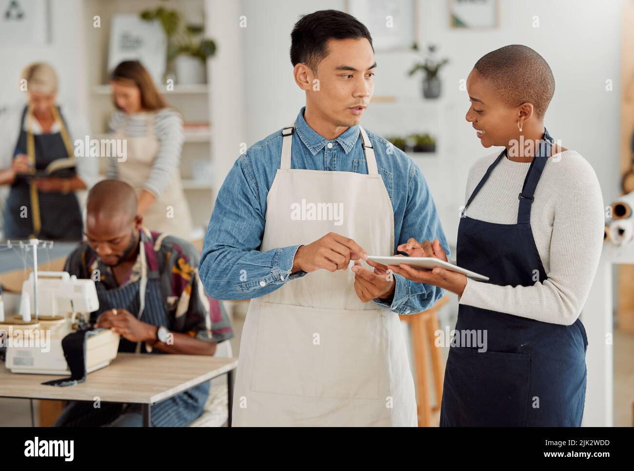 Unternehmer planen und diskutieren die Buchung von Nähkursen auf einem digitalen Tablet im Klassenzimmer. Nählehrer und Assistent sprechen darüber Stockfoto