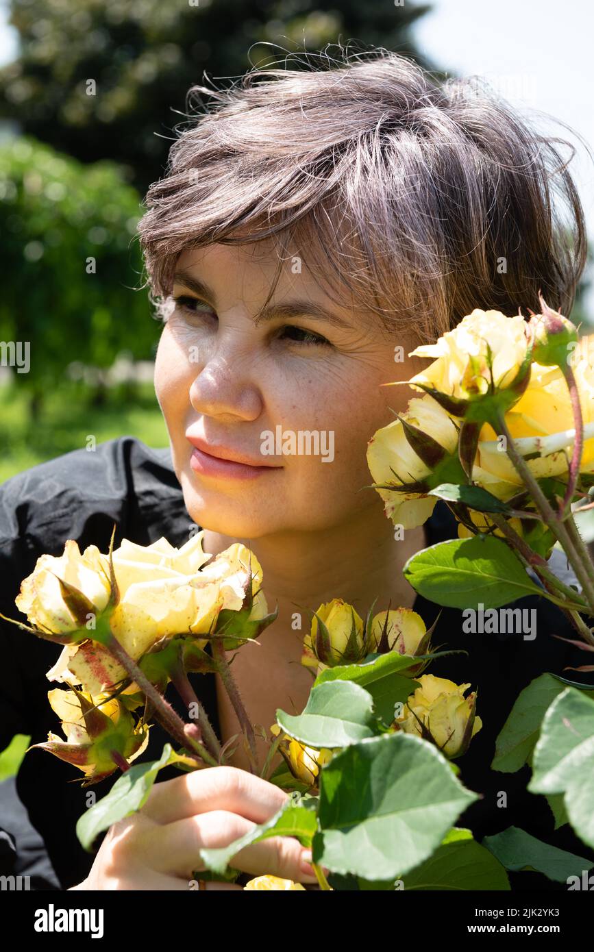 Frau mit einem niedlichen Lächeln schaut weg, während sie gelbe Rosen im Park wachsen hält. Stockfoto