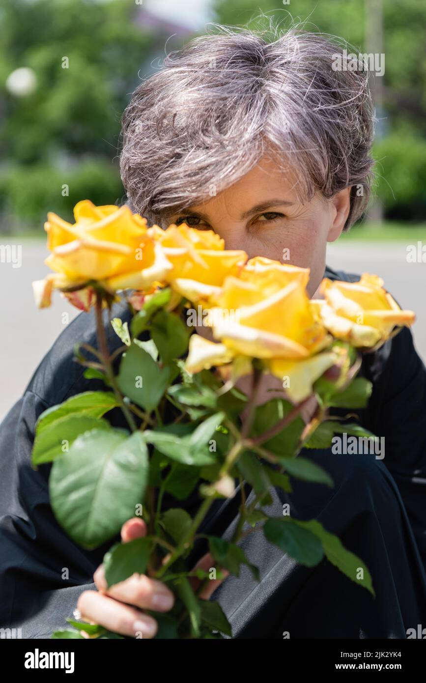 Ein Strauß blühender, gelber Rosen von hinten, von der eine Frau mit Interesse in ihren Augen auf die Kamera schaut. Stockfoto