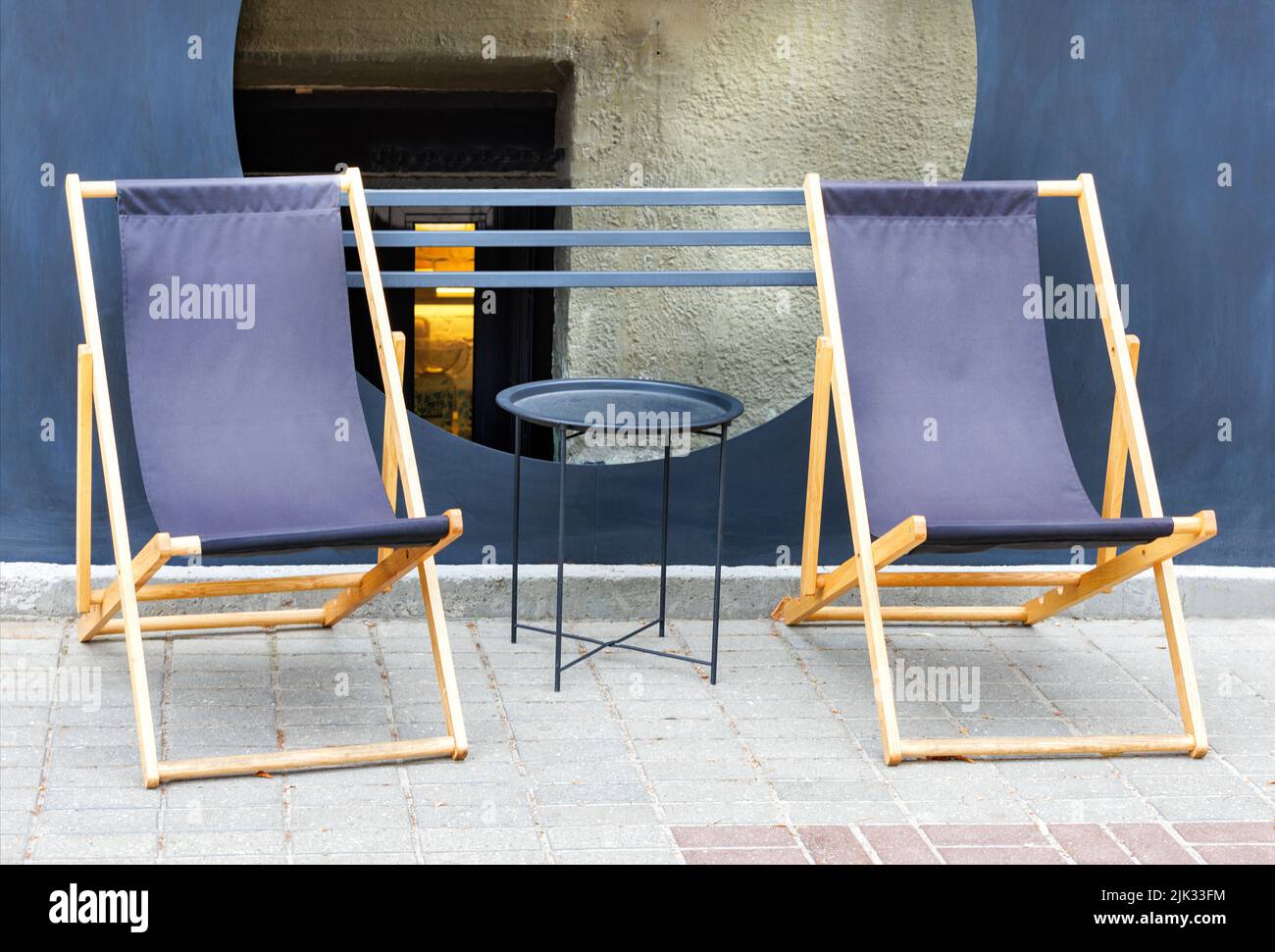 Zwei blaue Textil-Klappstühle mit einem Holzrahmen um einen Metalltisch vor dem Hintergrund eines gepflasterten Bürgerwegs und eines Café im Freien. Speicherplatz kopieren. Stockfoto