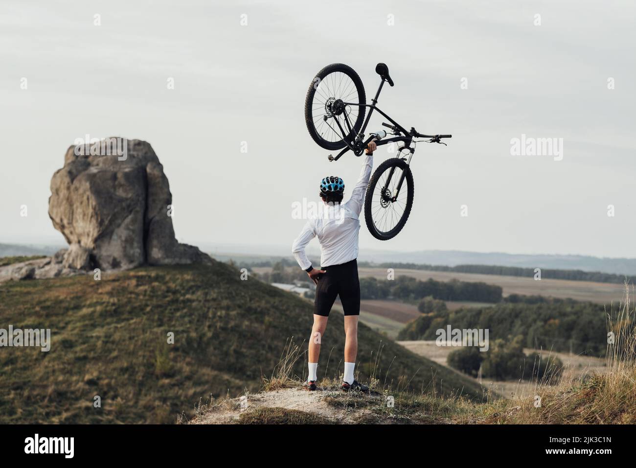 Professioneller Radsportler im Helm, der die Hand mit dem Fahrrad hebt, während er auf dem Hügel steht, riesiger Stein, Wunder der Natur auf dem Hintergrund Stockfoto