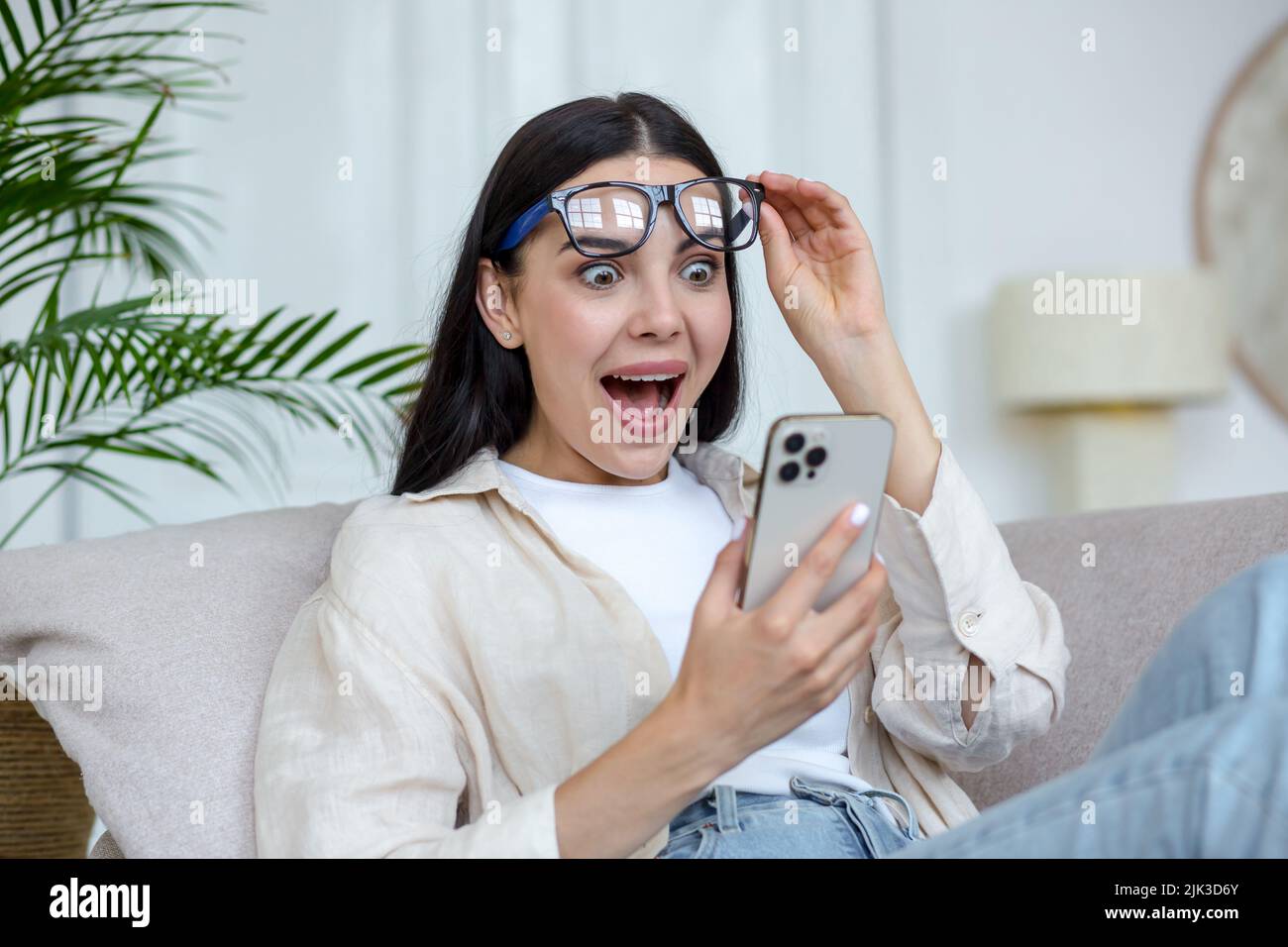 Junge schöne Frau in Brille freudig lesen Nachricht vom Handy, Brünette zu Hause auf dem Sofa sitzend ruhen Stockfoto