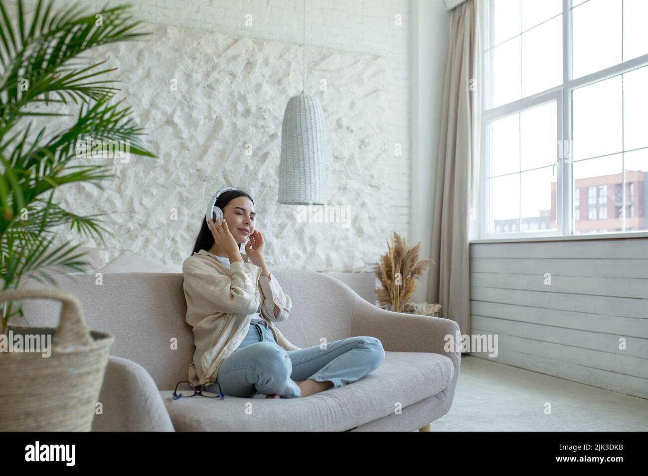 Schöne junge Frau, die sich ausruht, Musik in weißen großen Kopfhörern zu hören, Brünette, die im Wohnzimmer auf dem Sofa sitzt und mit geschlossenen Augen zu Hause träumt Stockfoto