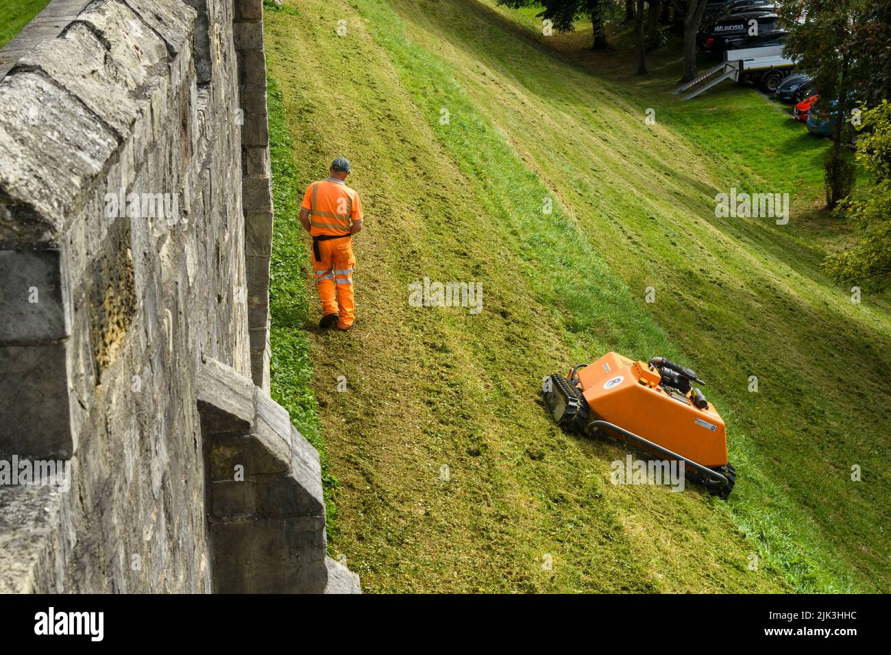Grashang, der von einem ferngesteuerten orangefarbenen Mähroboter (KommTek RoboFlail) und Arbeiter in der historischen Stadtmauer von York, Yorkshire, England, Großbritannien, gemäht wird. Stockfoto