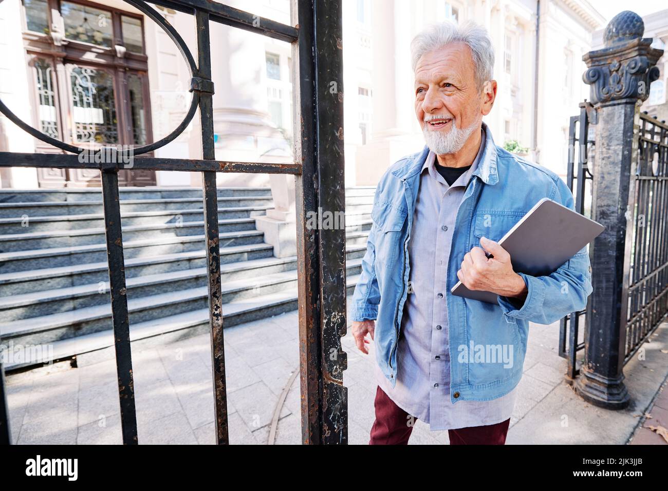 Ein glücklicher Seniorstudent des Umschulungsprogramms steht mit einem Laptop und Kaffee in den Händen vor der Universität. Es ist nie zu spät zu haben Stockfoto