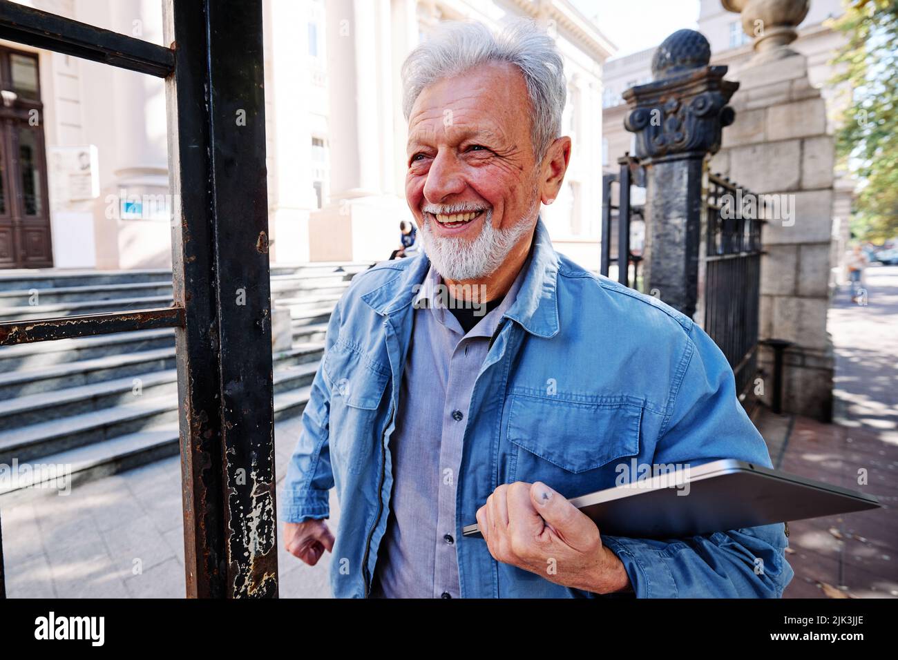 Ein glücklicher Seniorschüler verlässt die Schule. Er hält einen Laptop in der Hand. Umschulung für Senioren Konzept. Stockfoto