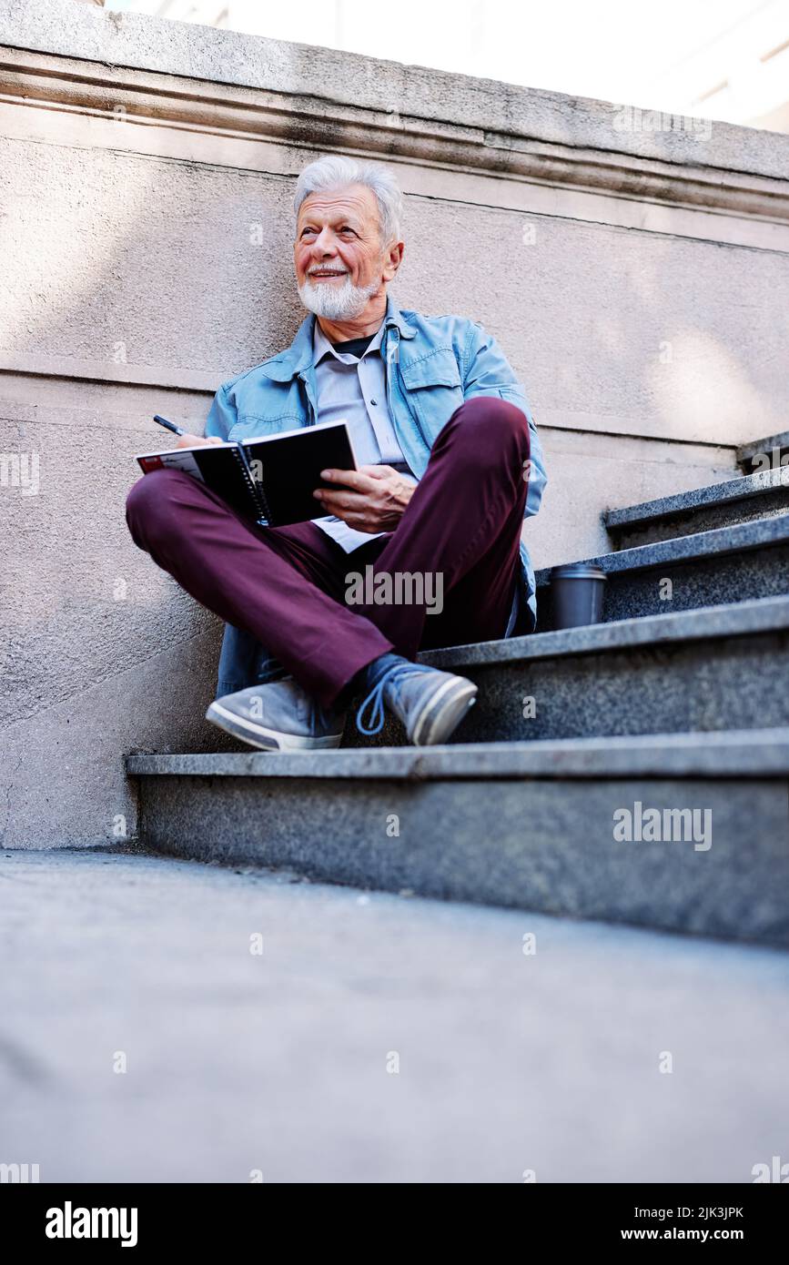 Ein kluger Senior Student, der auf der Treppe vor dem Universitätsgebäude sitzt und Hausaufgaben aufschreibt. Senioren in einem Umschulungsprogramm. Stockfoto
