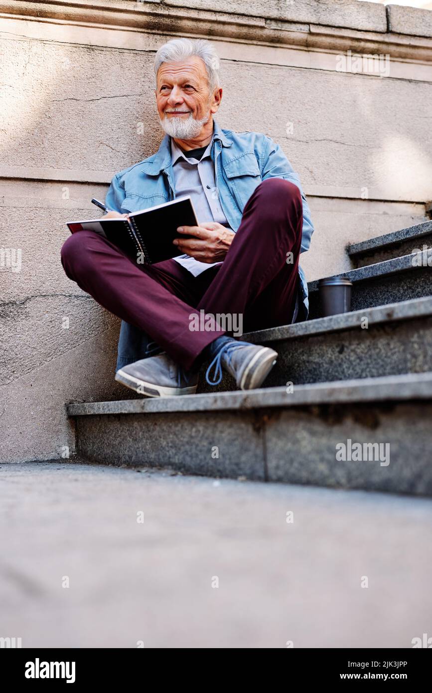 Ein glücklicher älterer Student sitzt auf der Treppe vor dem Universitätsgebäude und schreibt die Hausaufgaben auf. Umschulungsprogramm für Senioren. Stockfoto