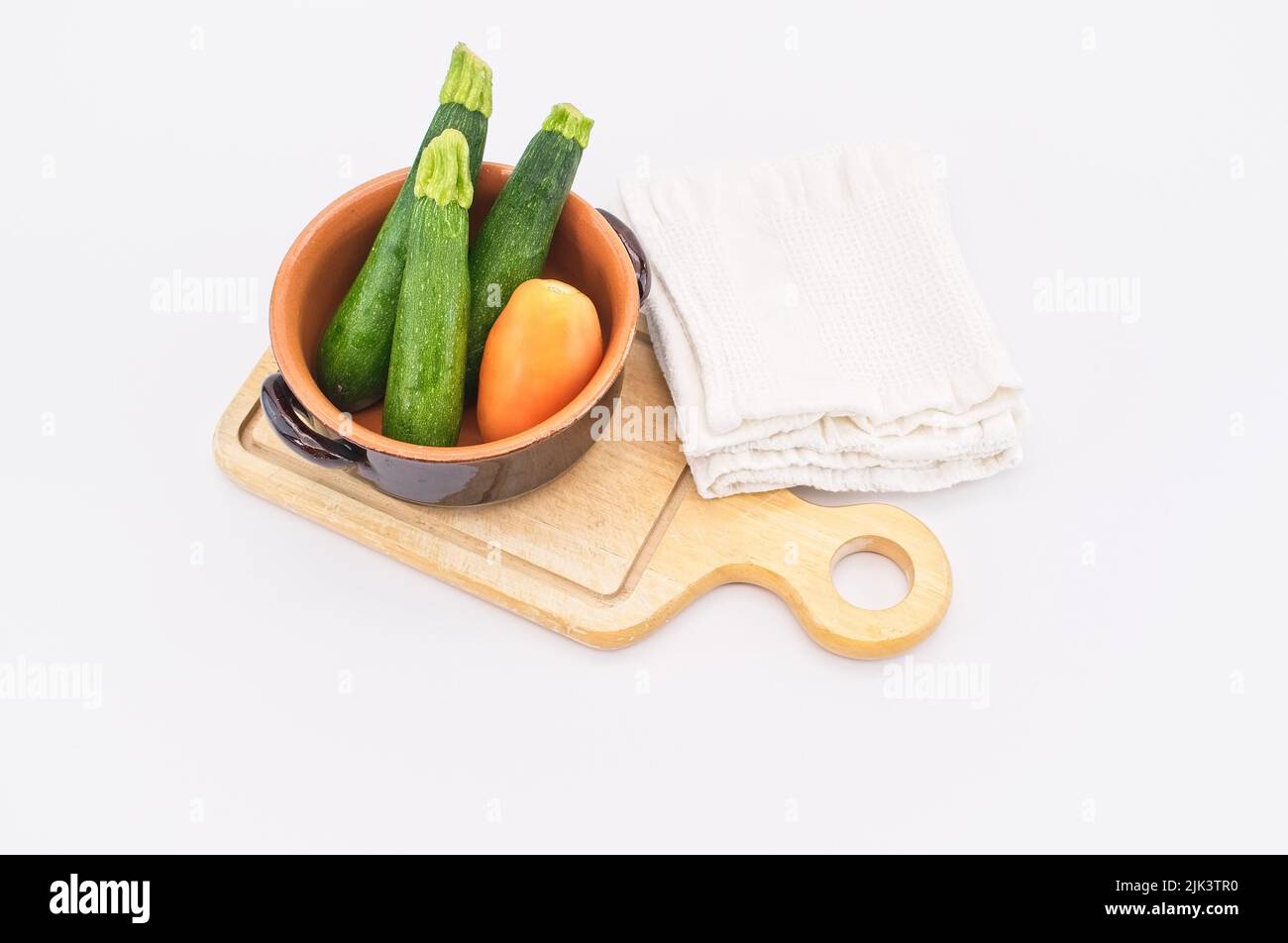 Frische Bio-Tomaten und Zucchini in einer Terrakotta-Schüssel mit Holzschneidebrett und Tuch auf einem weißen Tisch Stockfoto