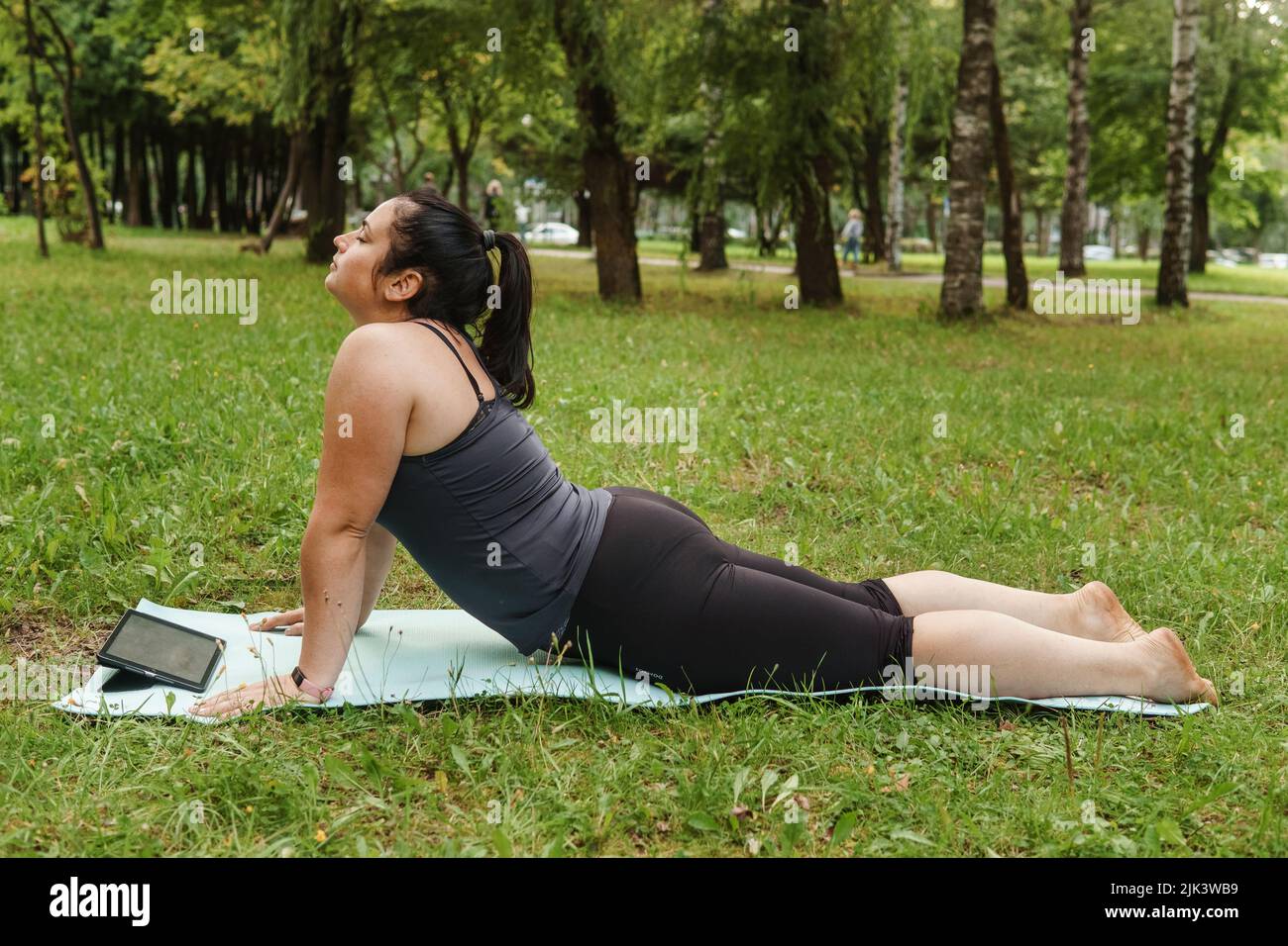 Eine charmante Brünette Frau plus-Größe Körper positiv praktiziert Sport in der Natur. Frau macht Yoga im Park auf einer Sportmatte. Auf einer Yogamatte im Hundepo Stockfoto