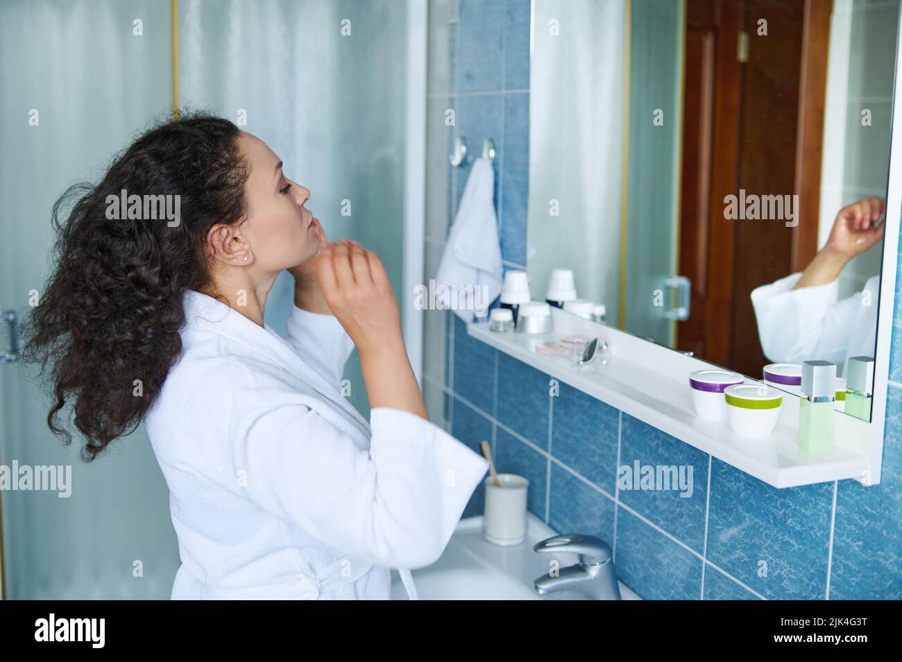 Hispanische Frau im weißen Bademantel, die im Badezimmer stand, spiegelte sich im Spiegel wider und berührte während der morgendlichen Routinen lockiges Haar Stockfoto