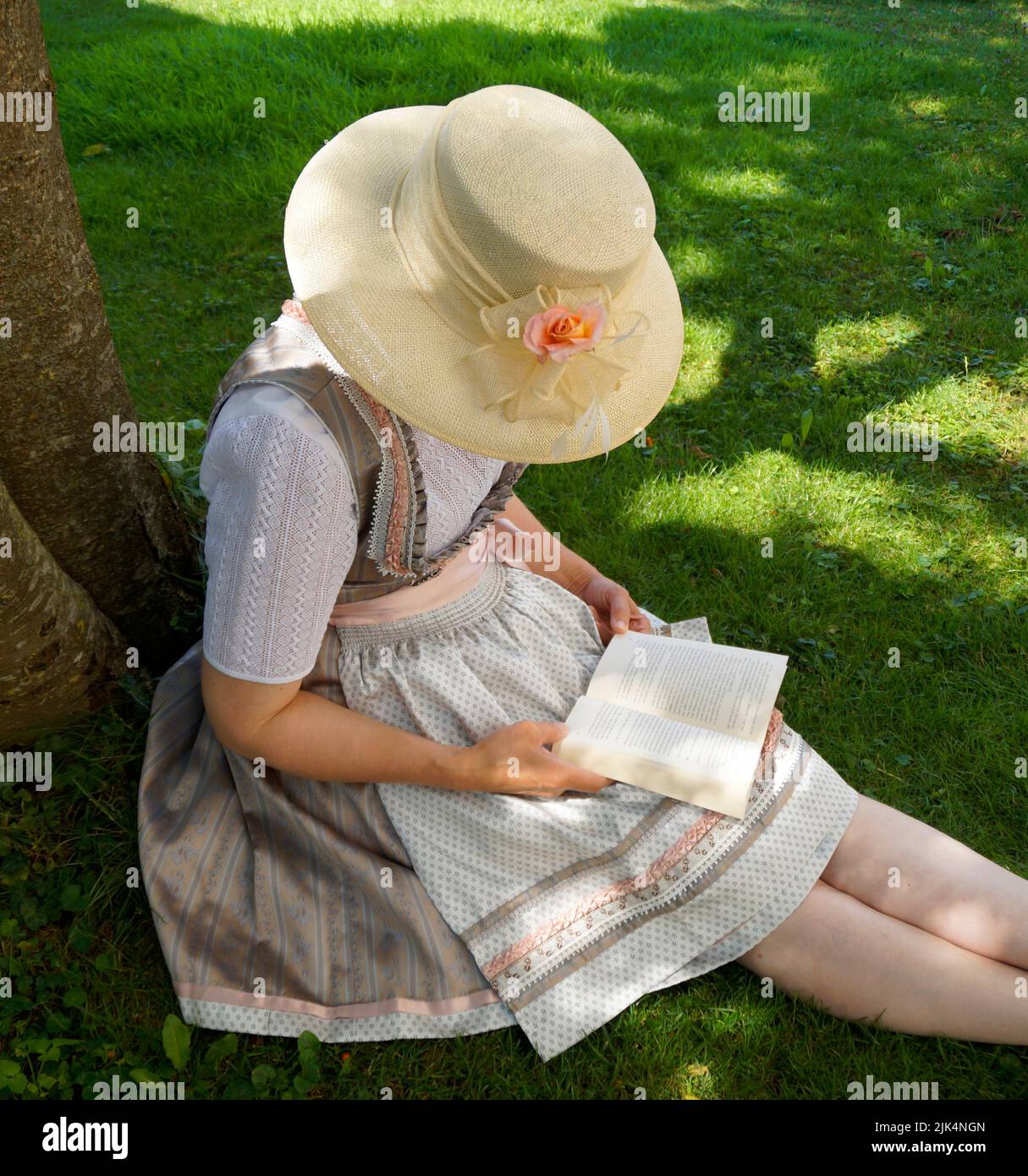 Eine Frau in einem schönen traditionellen bayerischen Dirndl-Kleid (oder Tracht), die an einem sonnigen Sommertag ein Buch auf der grünen Wiese liest (München, Bayern, Deutsch Stockfoto
