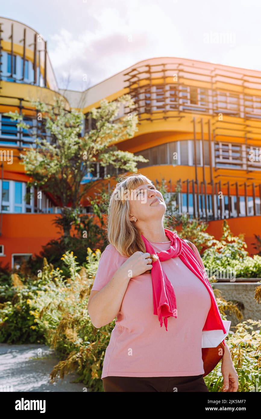 40s-jährige blonde Frau mit langen Haaren, in rosa Schal und Sommerkleidung, blickt auf moderne Architektur mit Hoffnung und Träumlichkeit. Konzept der housi Stockfoto
