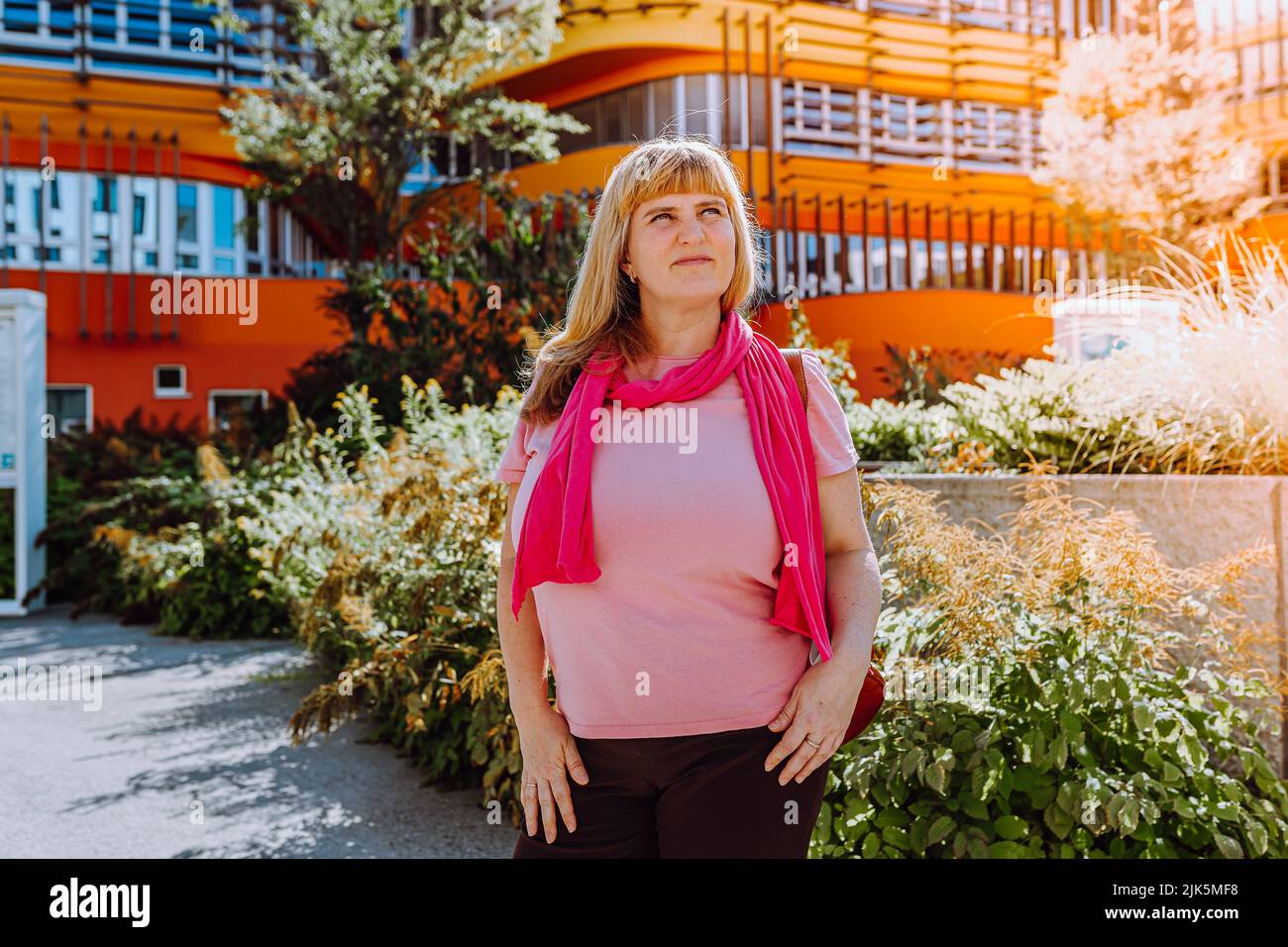 40s-jährige blonde Frau mit langen Haaren, in rosa Schal und Sommerkleidung, blickt auf moderne Architektur mit Hoffnung und Träumlichkeit. Konzept der housi Stockfoto