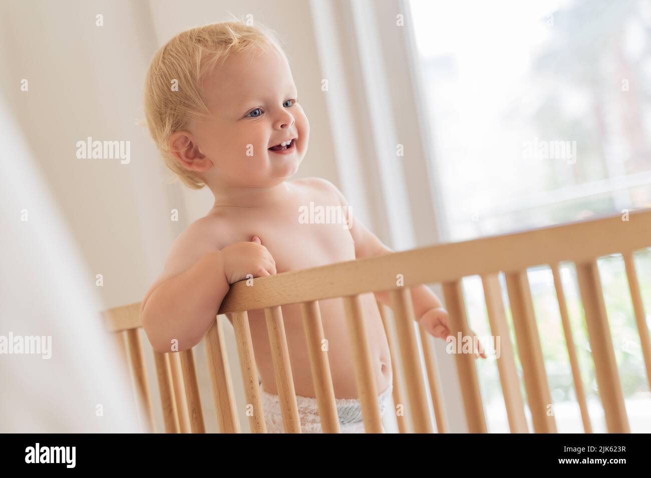 Indoor-Porträt von süßen kleinen Baby in Windel im Bett stehend versuchen zu gehen, lernen, den ersten Schritt halten an Stoßstangen auf Fensterhintergrund zu Hause zu tun Stockfoto