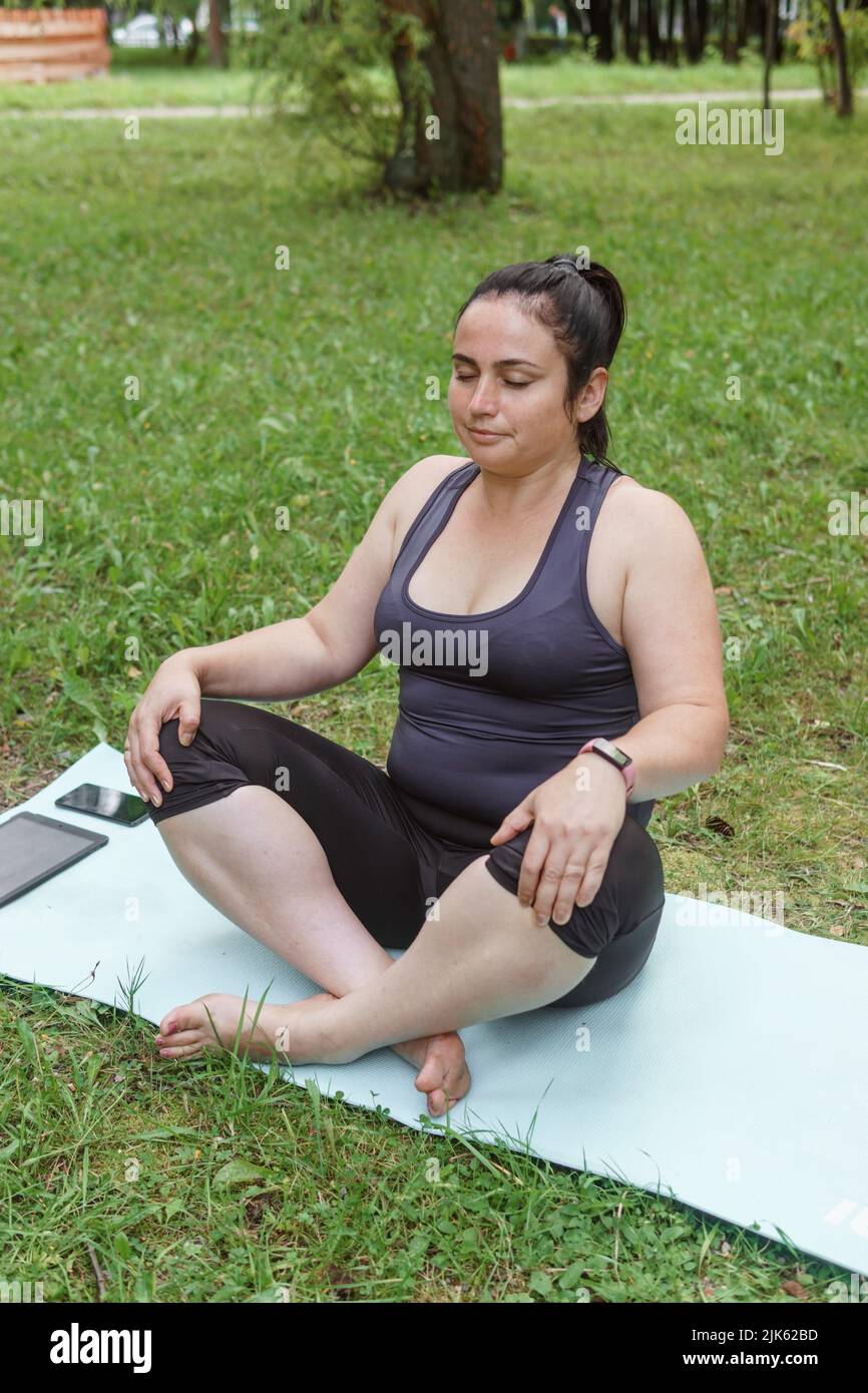 Eine charmante Brünette Frau plus-Größe Körper positiv praktiziert Sport in der Natur. Frau macht Yoga und Meditation im Park auf einer Sportmatte Stockfoto