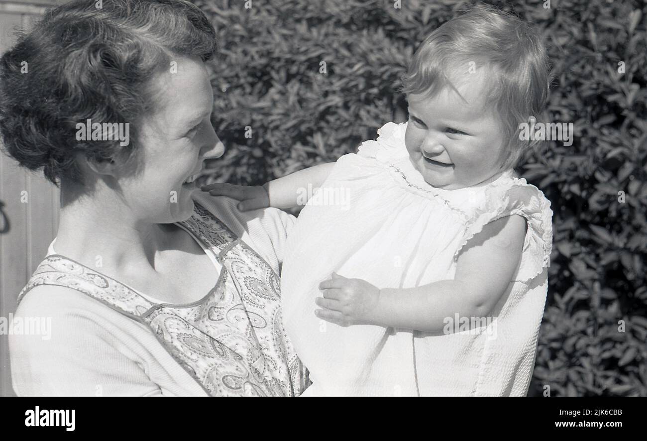 1960s, historisch, draußen im Vorgarten des Hauses, eine Mutter, die eine gemusterte Schürze der Zeit trug, ihre kleine Tochter in der Hand hielt und sie liebevoll ansah, England, Großbritannien. Stockfoto