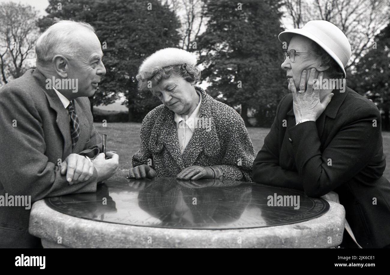 1960s, historisch, draußen in einem Park, drei gut gekleidete ältere Menschen, die auf dem Messinggesicht einer steinernen Sonnenuhr gelehnt sind, plaudern, England, Großbritannien. Stockfoto