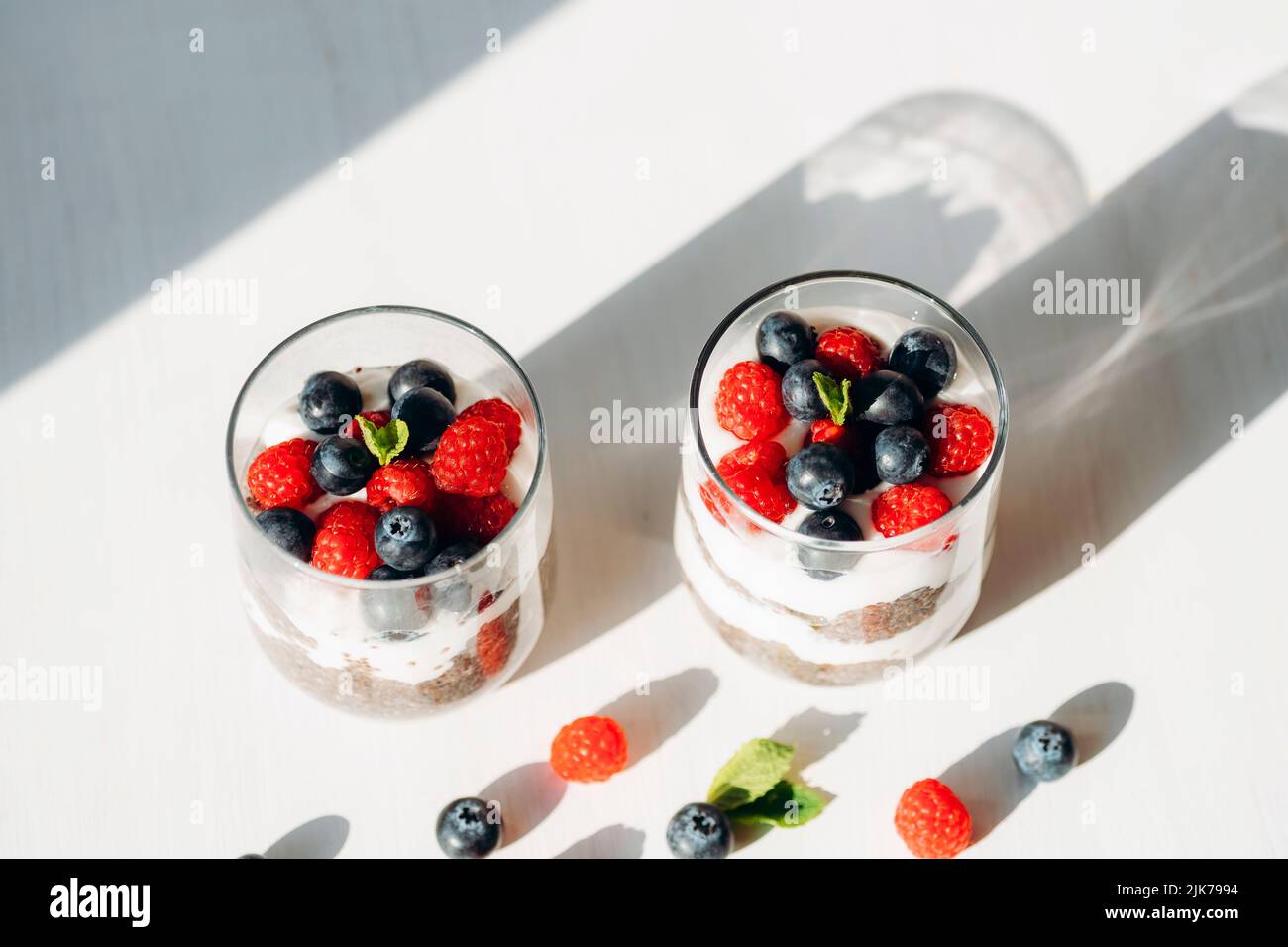 Zwei Gläser Chia Pudding mit frischer Himbeere und Heidelbeere auf weißem Tisch. Lange Schatten, Draufsicht. Stockfoto