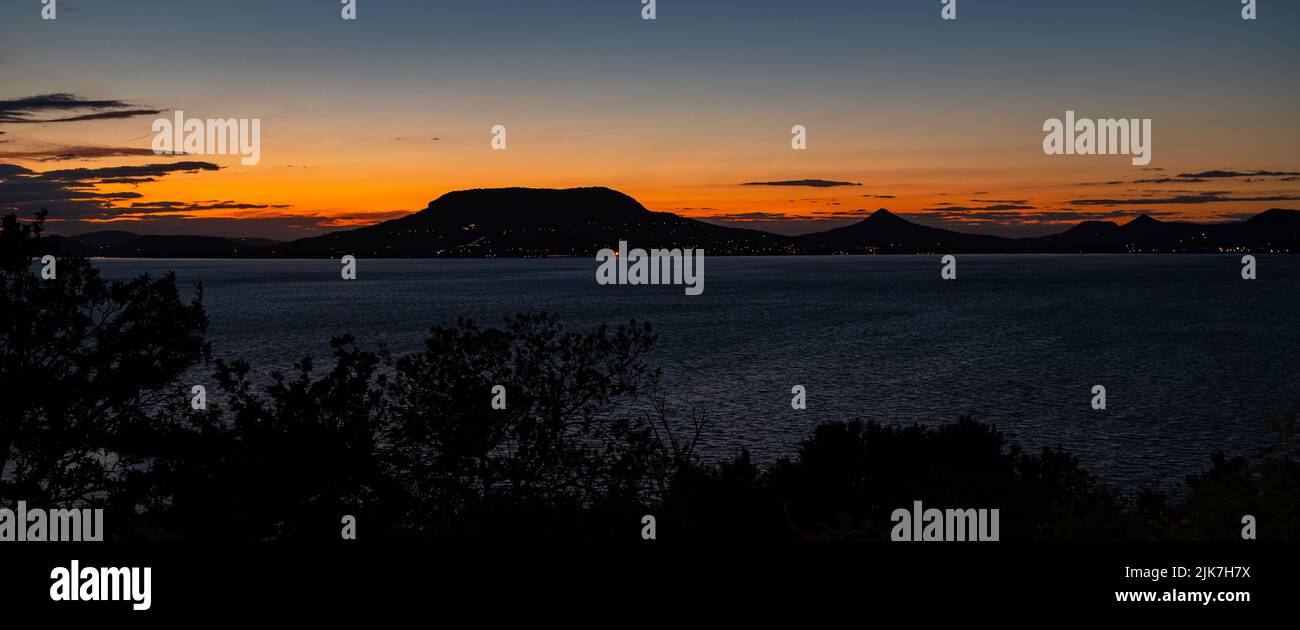 Panoramablick auf den Plattensee in Ungarn nach Sonnenuntergang. Hügel auf der anderen Seite des Sees ragen scharf gegen den bunten Dämmerungshimmel aus. Stockfoto
